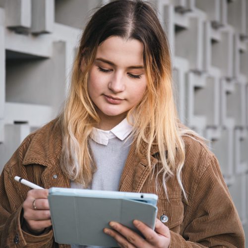 Attractive young woman in a corduroy jacket draws with a pencil on a digital tablet, on a blurred background of a geometric concrete wall in the city..